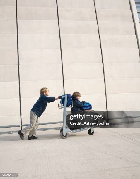 one boy pushing another on luggage trolley - ashley jouhar imagens e fotografias de stock