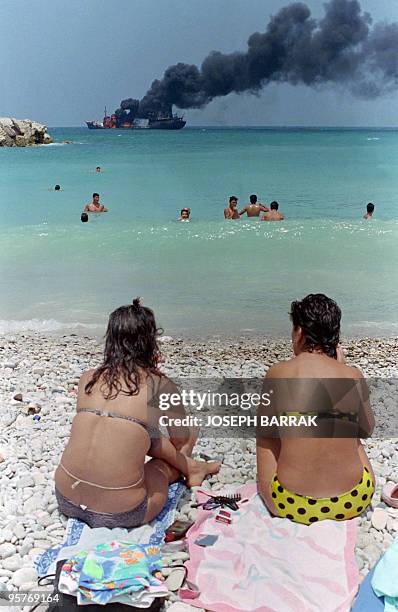 Two girls getting sun tan while the Chemicoral tanker still burns 16 June 1989 in front of Christian port of Jounieh in Christian eastern sector of...