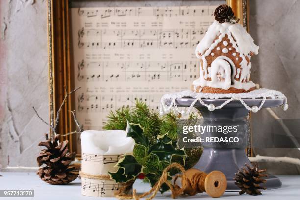 Small hand made Gingerbread house, candle and festive Christmas decor with vintage music notes over white table with plastered wall as background.