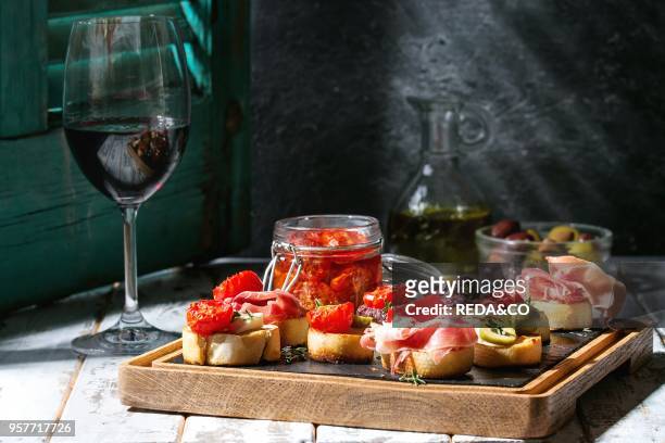 Tapas or bruschetta variety. Bread with ham prosciutto, sun dried tomatoes, olive oil, olives, pepper on slate wood board with glass of red wine over...