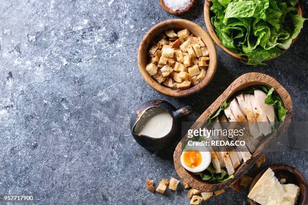 Bowls of ingredients for cooking classic Caesar salad. Sliced baked chicken breast, green roman salad, parmesan cheese, egg, croutons, salt, jug of...