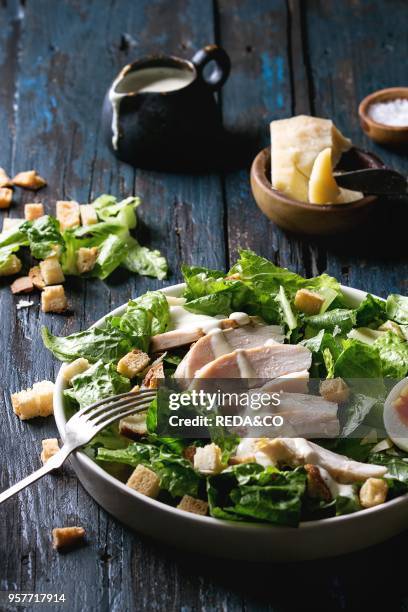 Classic Caesar salad with grilled chicken breast and half of egg in white ceramic plate. Served with fork and ingredients above over old dark blue...