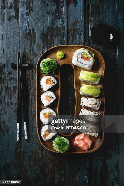 Homemade sushi rolls set with salmon, sesame seeds and avocado serving in wood plate with pink pickled ginger, soy sauce, wasabi, seaweed salad,...