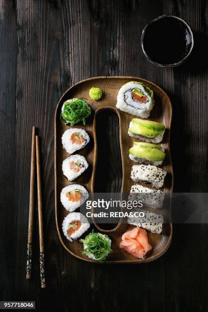 Homemade sushi rolls set with salmon, sesame seeds and avocado serving in wood plate with pink pickled ginger, soy sauce, wasabi, seaweed salad,...