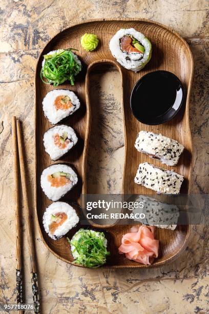 Homemade sushi rolls set with salmon, sesame seeds serving in wood plate with pink pickled ginger, soy sauce, wasabi, seaweed salad, chopsticks over...