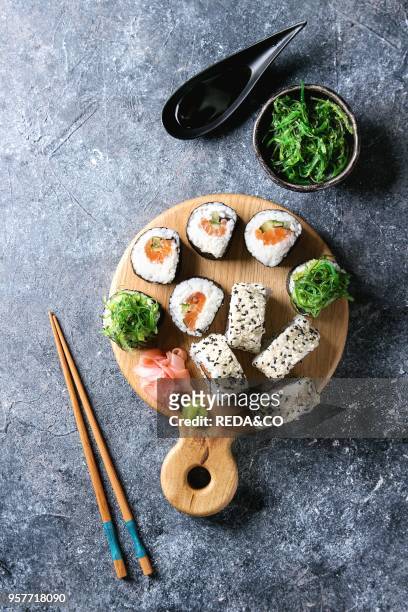 Homemade sushi rolls set with salmon, sesame seeds serving on wood serving board with pink pickled ginger, soy sauce, wasabi, seaweed salad,...