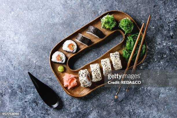 Homemade sushi rolls set with salmon, sesame seeds serving in wooden plate with pink pickled ginger, soy sauce, wasabi, seaweed salad, chopsticks on...