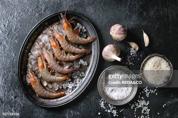 Raw whole fresh uncooked prawns shrimps on ice on vintage metal tray with cooking ingredients above. Sea salt, rice, garlic, over black texture...