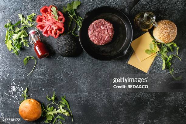 Ingredients for cooking hamburger. Meat beef burger in pan, cheese, ketchup sauce, tomato, black and white buns, arugula salad over dark texture...