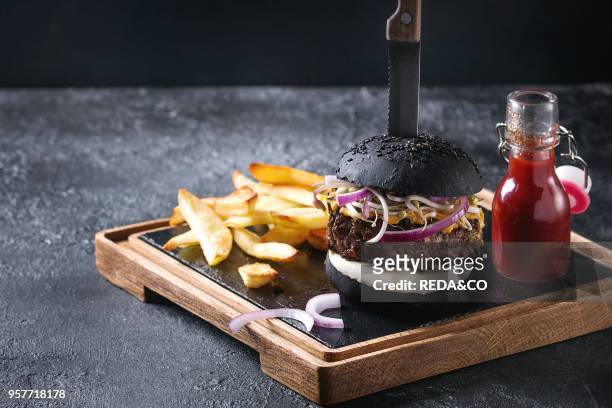 Homemade black bun hamburger with beef, mozzarella cheese, sprouts served on wooden slate serving board with french fries, knife and ketchup sauce on...