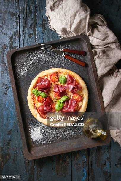 Traditional pizza with bresaola, cheese, tomatoes and basil served on old oven tray with textile over dark blue wooden background. Top view with...