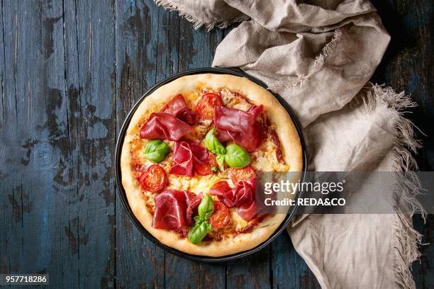 Traditional pizza with bresaola, cheese, tomatoes and basil served on black plate with textile over dark blue wooden background. Top view with space....