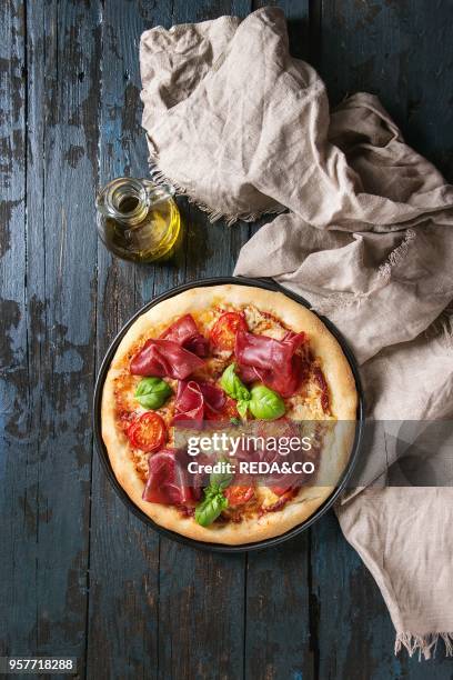 Traditional pizza with bresaola, cheese, tomatoes and basil served on black plate with textile and olive oil over dark blue wooden background. Top...