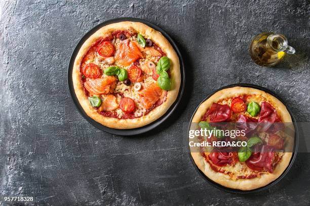 Two Traditional pizza with bresaola, smoked salmon, cheese, tomatoes and basil served on black plates with olive oil over black texture background....