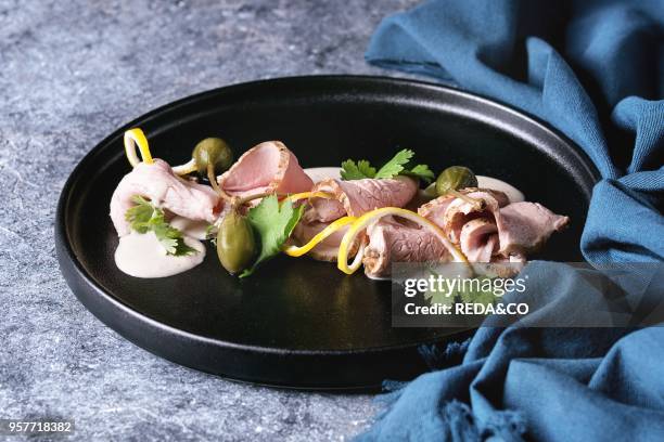 Vitello tonnato italian dish. Thin sliced veal with tuna sauce, capers and coriander served on black plate with blue textile over blue texture...