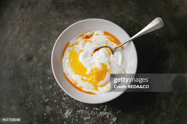 Poached egg on yogurt sauce with spicy olive oil, salt, spoon served in white plate over old dark metal background. Top view, copy space. Vegetarian...