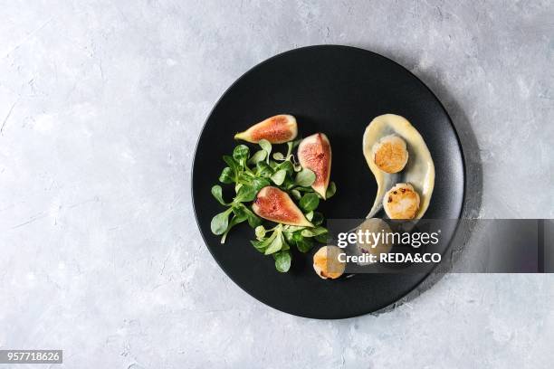 Fried scallops with lemon, figs, sauce and green salad served on black plate over gray texture background. Top view, copy space. Plating, fine dining.