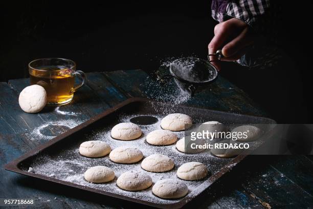 Homemade almond cookies on old oven tray and cup of hot tea over dark blue wooden table. Sprinkling sugar powder by vintage sieve in child hand. Dark...