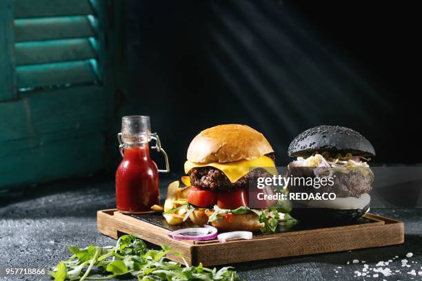 Homemade black and white buns hamburgers with beef, mozzarella cheese, sprouts, arugula, served on wooden slate serving board with french fries and...