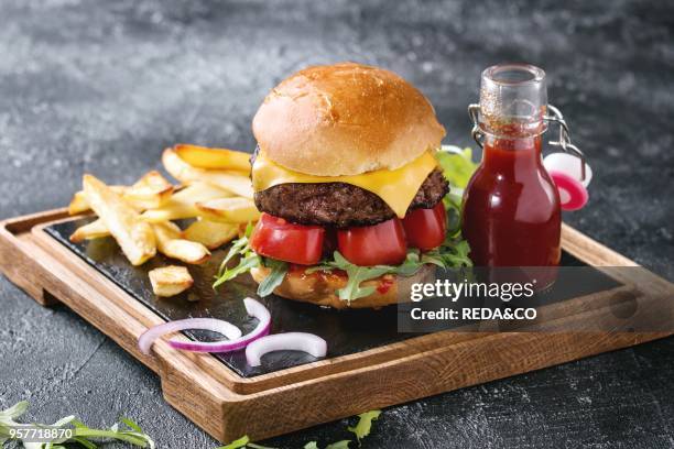 Homemade traditional hamburger with beef, tomato, cheese, aragula, served on wooden slate serving board with french fries and ketchup sauce on dark...