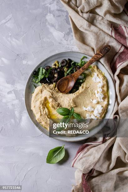 Homemade traditional spread hummus with chopping olives, oil and herbs on blue plate, served with feta cheese, spoon, basil, textile on gray texture...