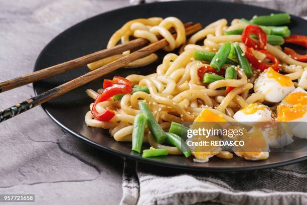Cooking stir fry udon noodles, green beans, sliced paprika, boiled eggs, soy sauce with sesame seeds in black plate with wood chopsticks over gray...