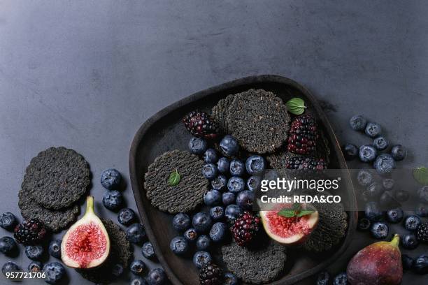 Variety of fresh berries blueberry, dewberry, red currant and figs with black charcoal crackers on wooden plate over dark metal background. Top view...
