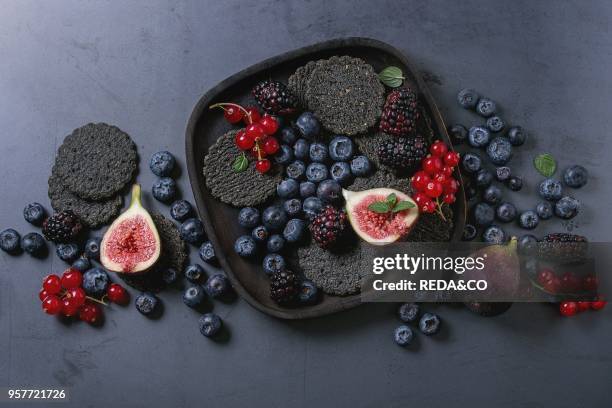 Variety of fresh berries blueberry, dewberry, red currant and figs with black charcoal crackers on wooden plate over dark metal background. Top view...