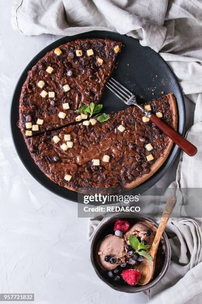 Dessert chocolate pizza with dark, milk, white chocolate, served on black plate with fork, mint, frozen berries and bowl of ice cream over gray...