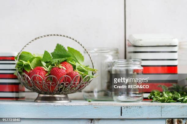 Fresh ripe garden strawberries and melissa herbs in vintage vase standing with empty glass and metal jars for jam on blue white wooden kitchen table....