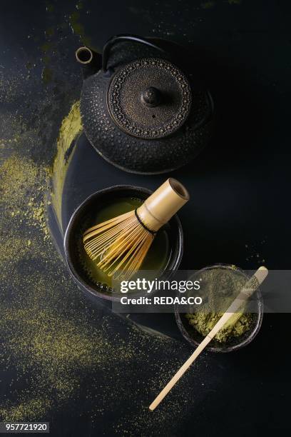 Green tea matcha powder and hot drink in black bowls standing with iron teapot, bamboo traditional tools spoon and whisk over dark metal background....