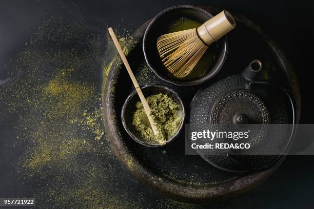 Green tea matcha powder and hot drink in black bowls standing with iron teapot, bamboo traditional tools spoon and whisk in terracotta tray over dark...