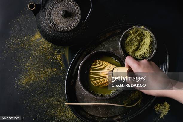 Green tea matcha powder and hot drink in black bowls standing with iron teapot, bamboo traditional tools spoon and whisk in hand in vintage tray over...