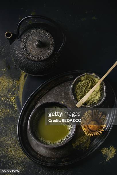 Green tea matcha powder and hot drink in black bowls standing with iron teapot, bamboo traditional tools spoon and whisk in vintage tray over dark...