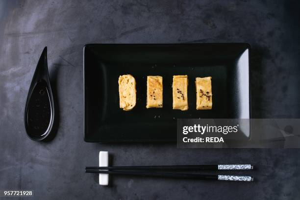 Japanese Rolled Omelette Tamagoyaki sliced with black seasame seeds and soy sauce, served in black square ceramic plate with chopsticks over dark...