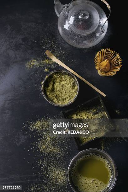 Green tea matcha powder and hot drink in black bowls and wood box standing with glass teapot, bamboo traditional tools spoon, whisk in terracotta...