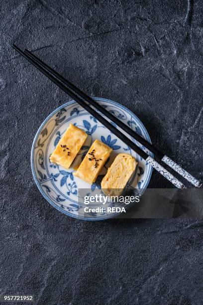 Japanese Rolled Omelet Tamagoyaki sliced with black sesame seeds and soy sauce, served in blue white ornate ceramic plate with chopsticks over black...
