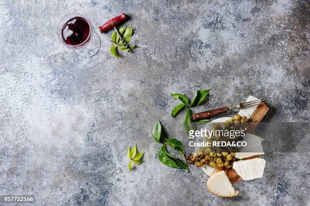 Serving board with sliced camembert cheese and baked bunch of green grapes served with bread, glass of red wine, corkscrew, green leaves, fork over...