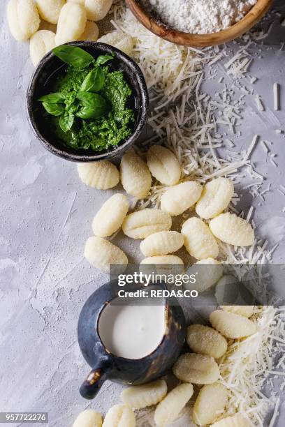 Raw uncooked potato gnocchi in black wooden plates with ingredients. Flour, grated parmesan cheese, basil pesto sauce, jug of cream over gray...