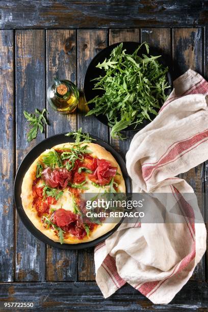 Whole homemade pizza with cheese and bresaola, served on black plate with fresh arugula, olive oil and kitchen towel over old wooden plank...