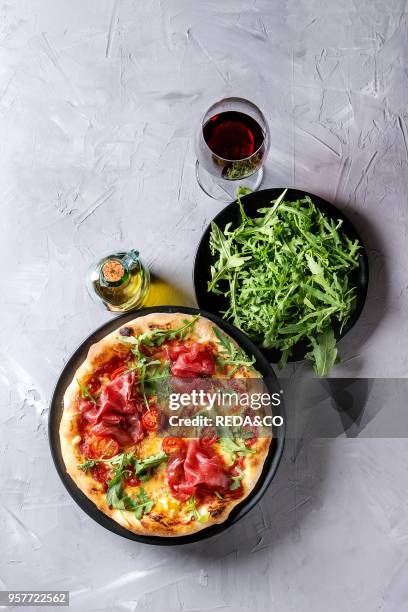 Whole homemade pizza with cheese and bresaola, served on black plate with fresh arugula, olive oil, glass of red wine over gray concrete texture...