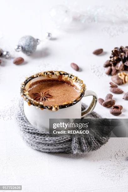 Vintage mug in wool scarf of hot chocolate, decor with nuts, caramel, spices. Ingredients and Christmas toys above over white texture background with...