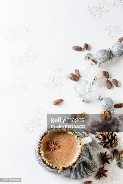 Vintage mug in wool scarf of hot chocolate, decor with nuts, caramel, spices. Ingredients and Christmas toys above over white texture background. Top...