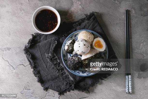 Blue plate with different size rice balls with black sesame and seaweed nori, served with soft boiled eggs, soy sauce, chopsticks over gray table....