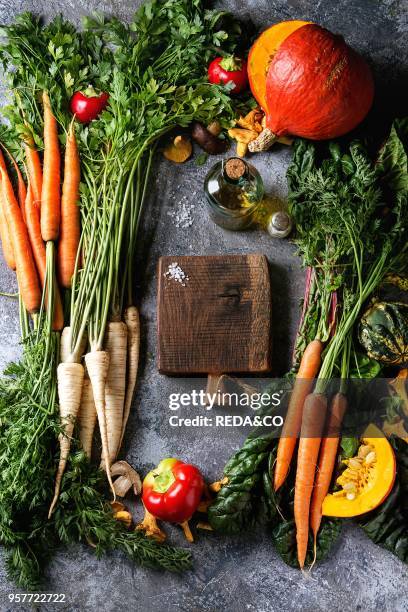 Variety of autumn harvest vegetables carrot, parsnip, chard, paprika, hokkaido pumpkin, mushrooms. Empty wooden chopping board, olive oil, salt over...