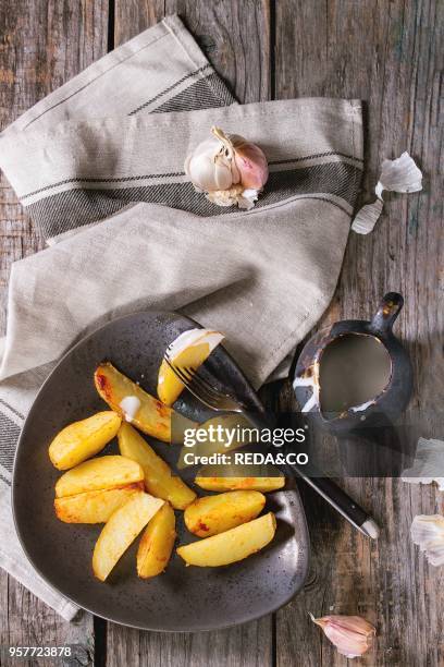 Fried country potatoes on brown ceramic plate, served with homemade aioli sauce in jug, garlic and vintage fork on gray linen napkin over wooden...