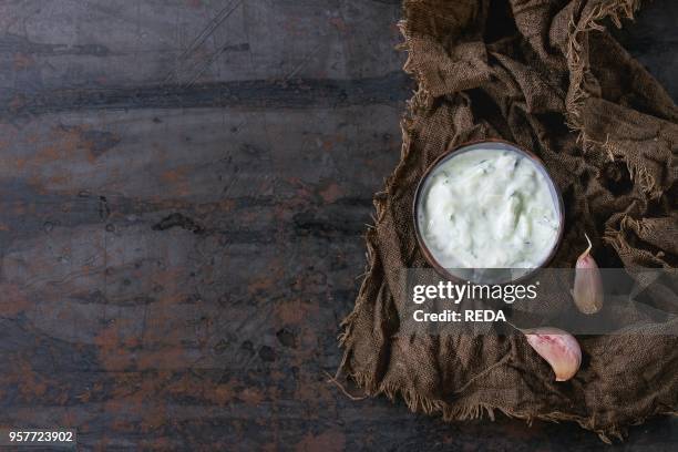 Ceramic bowl of traditional greek sauce tzatziki with cucumber and garlic, served on wet sackcloth rag over old dark textured iron background. Top...