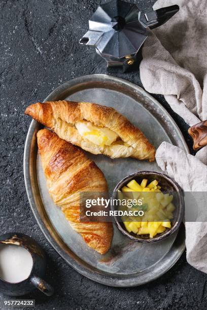 Breakfast with two croissant, butter, coffee maker, cream and sliced mango fruit, served on serving metal tray with textile napkin over black...