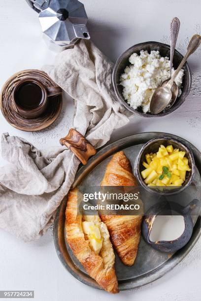 Breakfast with two croissant, butter, cup of coffee, cottage cheese and sliced mango fruit, served on serving metal tray with textile napkin on white...