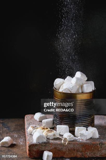 Heap of homemade vanilla marshmallow fresh and fried with sprinkling sugar powder in old tin can on terracotta board over dark wooden background....